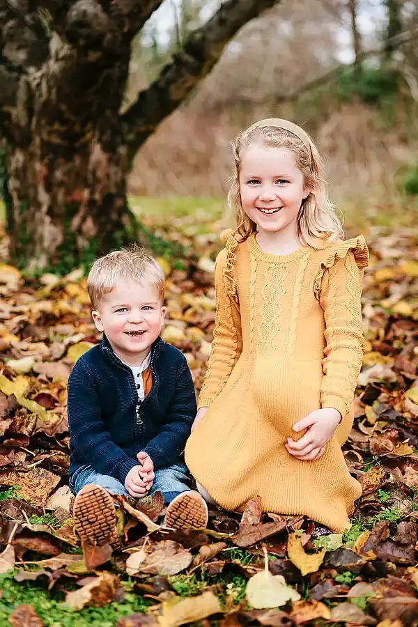 Brother and sister kneeling in the woods amongst autumnal leaves.